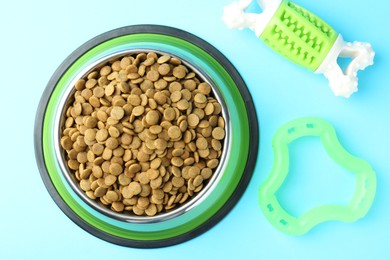 Photo of Dry pet food in feeding bowl and toys on light blue background, flat lay