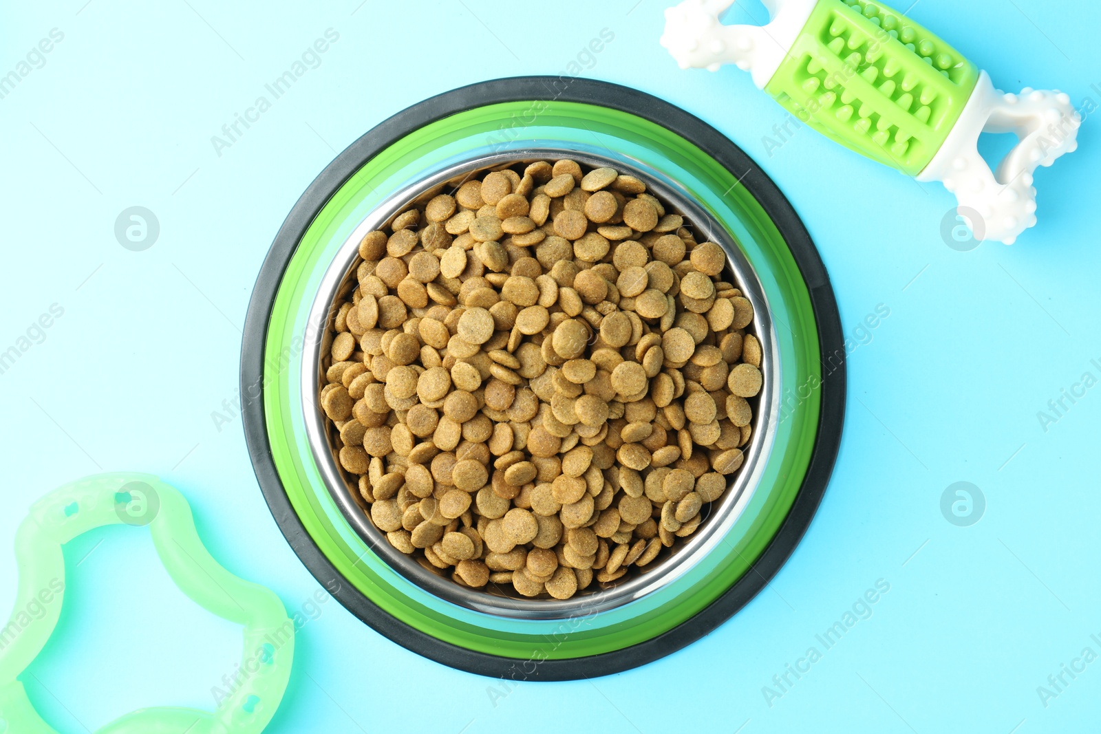 Photo of Dry pet food in feeding bowl and toys on light blue background, flat lay