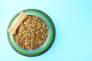 Photo of Dry pet food in feeding bowl and chew bone on light blue background, top view. Space for text