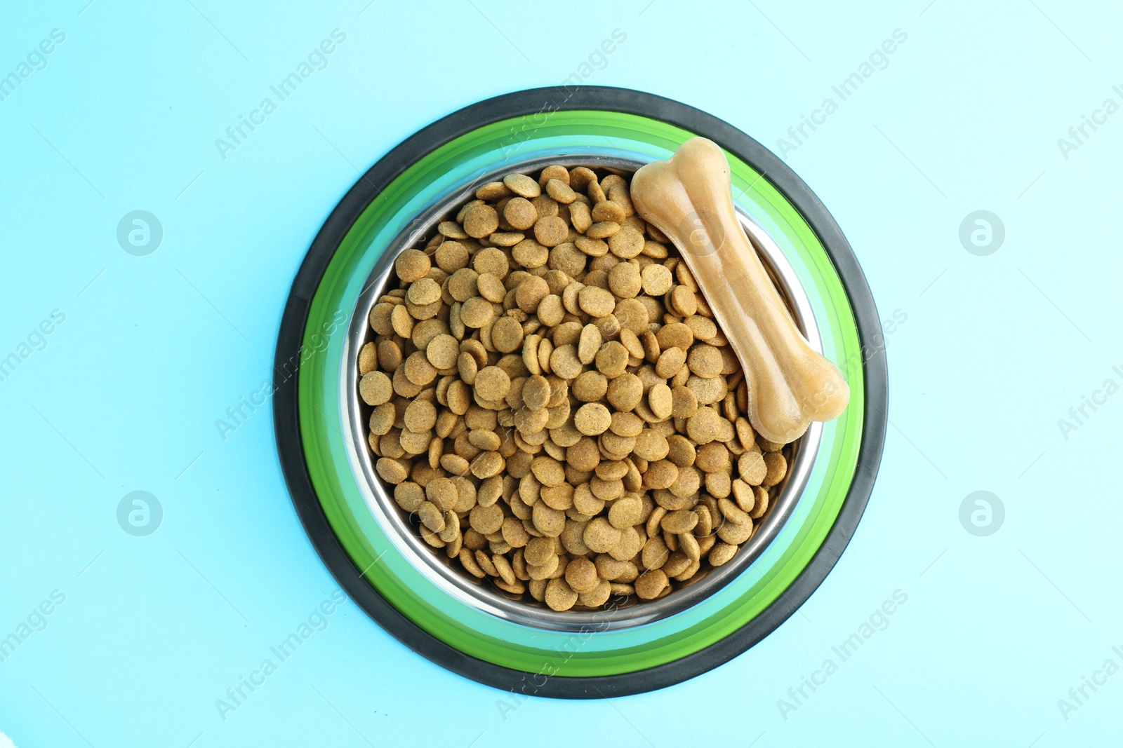 Photo of Dry pet food in feeding bowl and chew bone on light blue background, top view