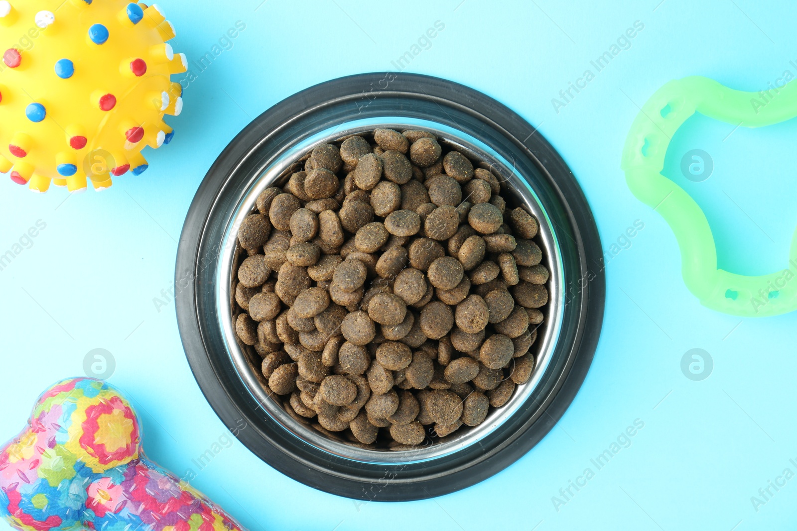 Photo of Dry pet food in feeding bowl and toys on light blue background, flat lay