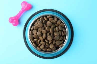 Photo of Dry pet food in feeding bowl and chew bone on light blue background, flat lay