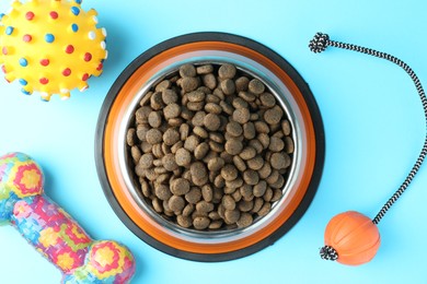 Photo of Dry pet food in feeding bowl and toys on light blue background, flat lay