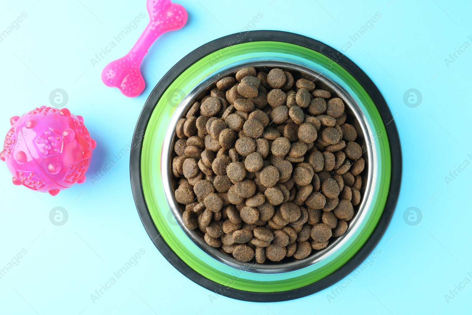 Photo of Dry pet food in feeding bowl and toys on light blue background, flat lay