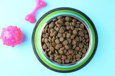 Photo of Dry pet food in feeding bowl and toys on light blue background, flat lay