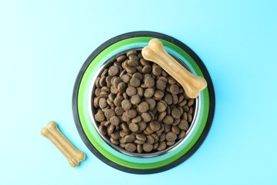 Photo of Dry pet food in feeding bowl and chew bones on light blue background, top view