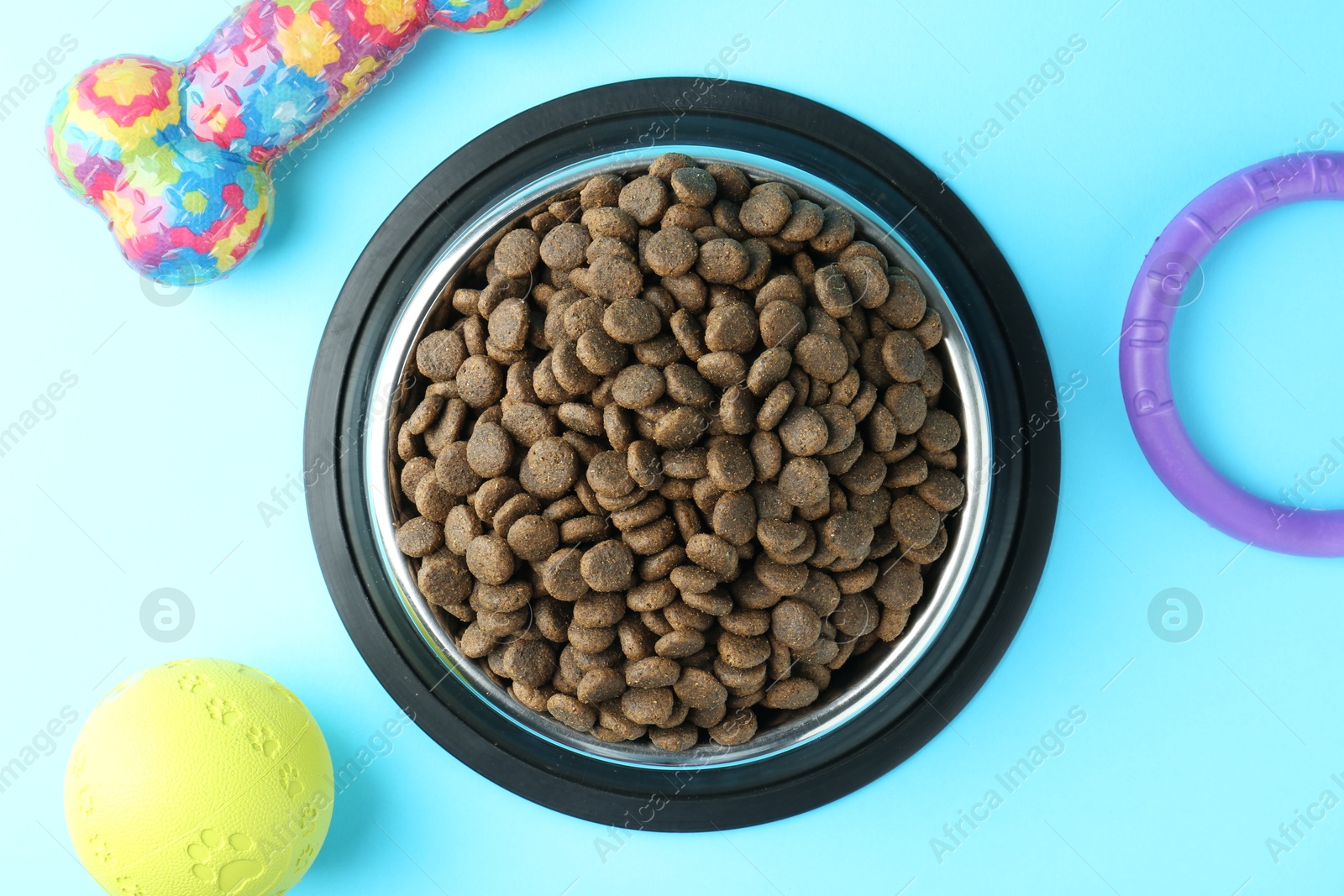 Photo of Dry pet food in feeding bowl and toys on light blue background, flat lay