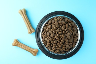 Photo of Dry pet food in feeding bowl and chew bones on light blue background, top view