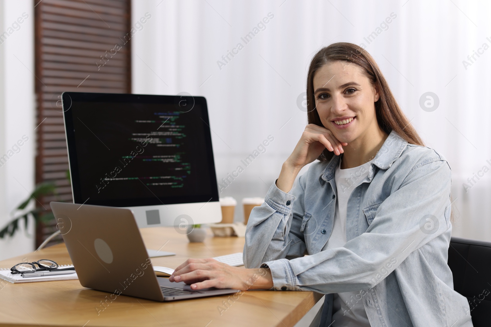 Photo of Programmer working on laptop and computer at wooden desk indoors