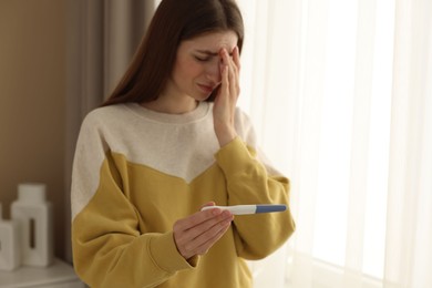 Upset woman with negative pregnancy test near window at home, space for text