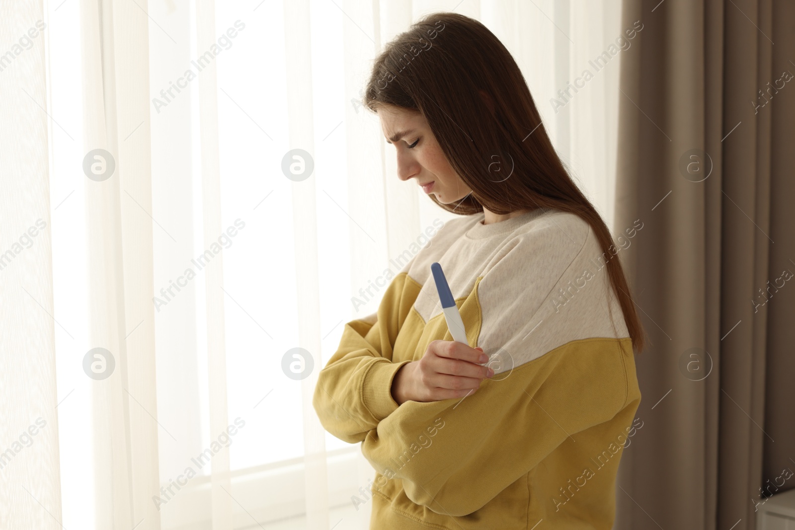 Photo of Upset woman with negative pregnancy test near window at home, space for text