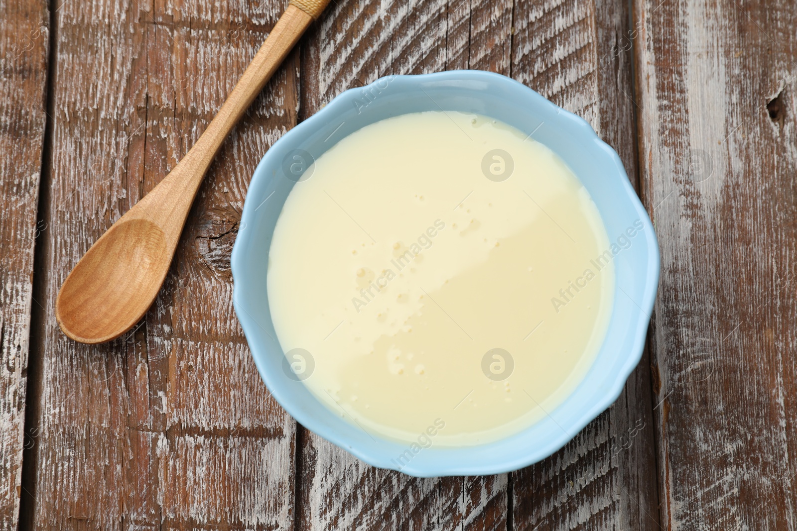 Photo of Condensed milk in bowl and spoon on wooden table, top view