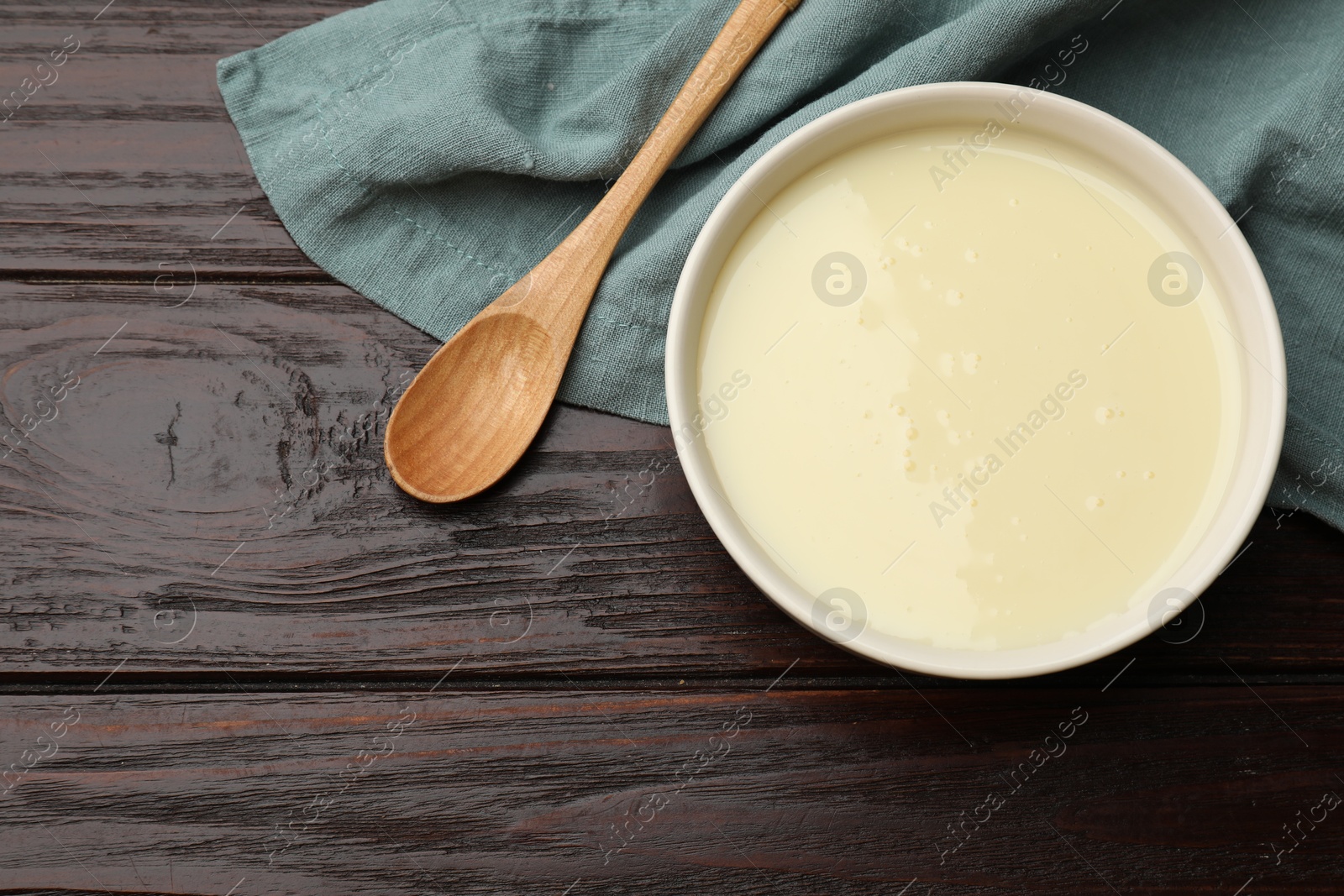 Photo of Condensed milk in bowl and spoon on wooden table, top view. Space for text