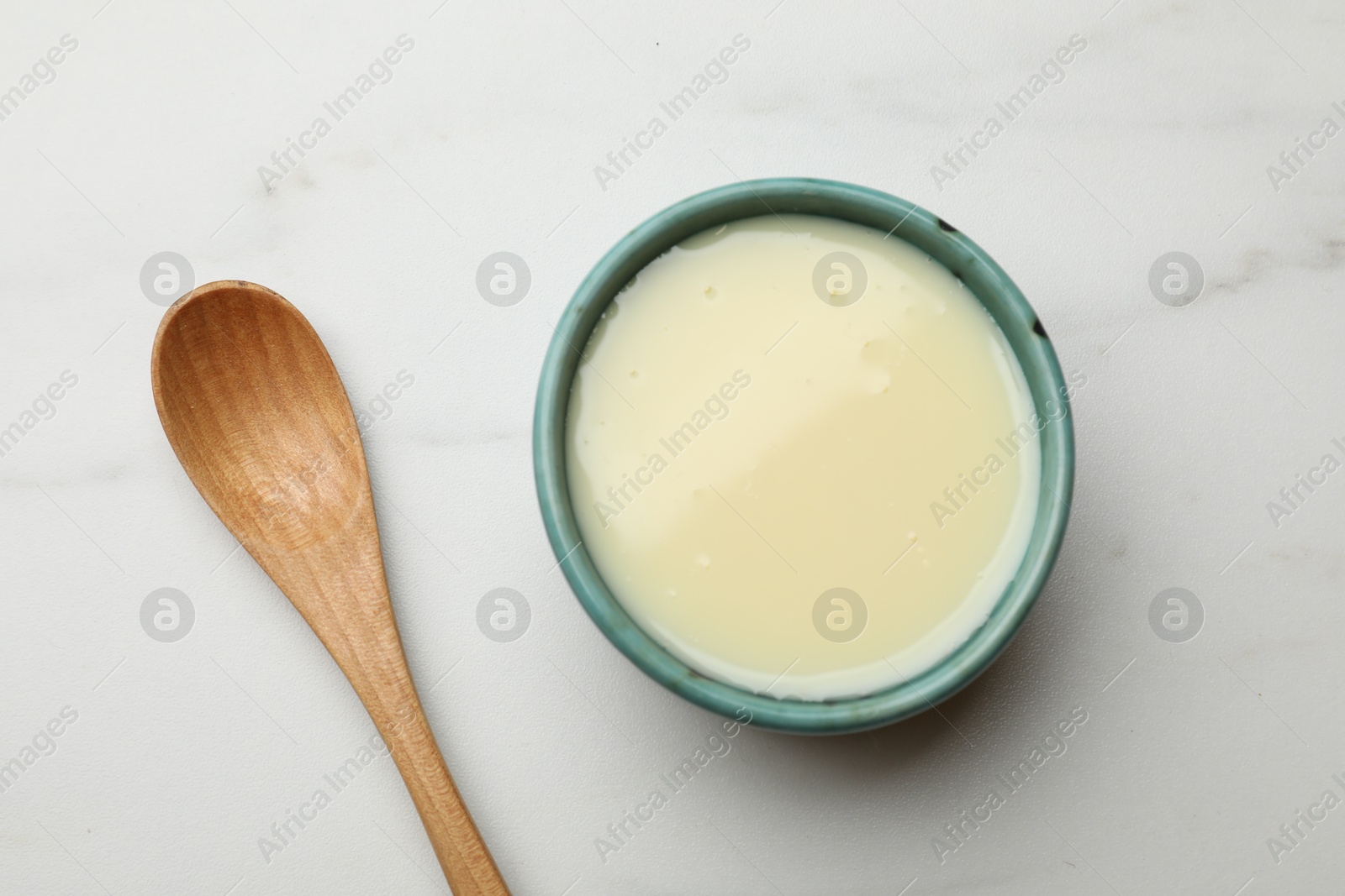 Photo of Condensed milk in bowl and spoon on white marble table, top view