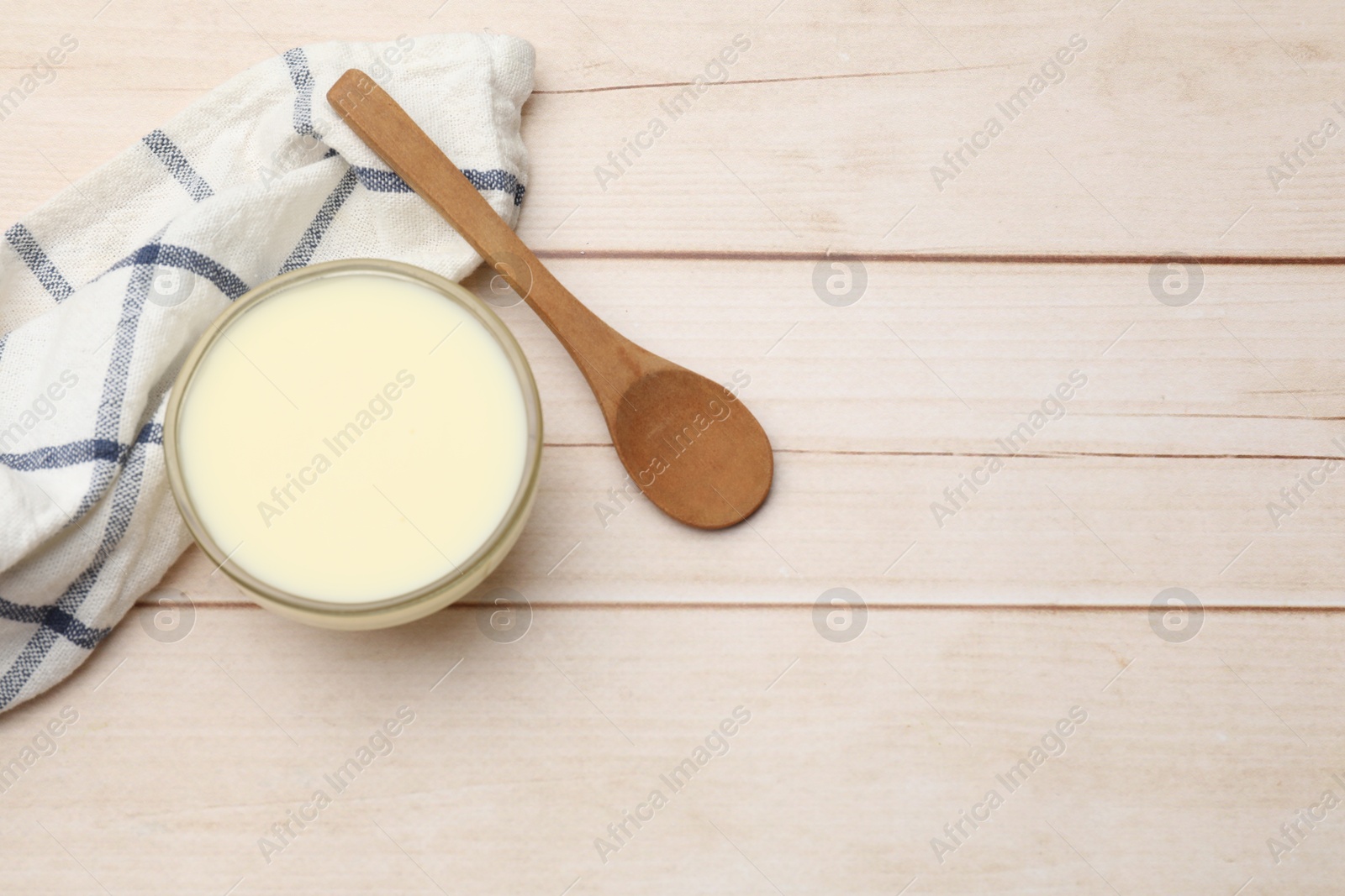 Photo of Condensed milk in bowl and spoon on light wooden table, top view. Space for text