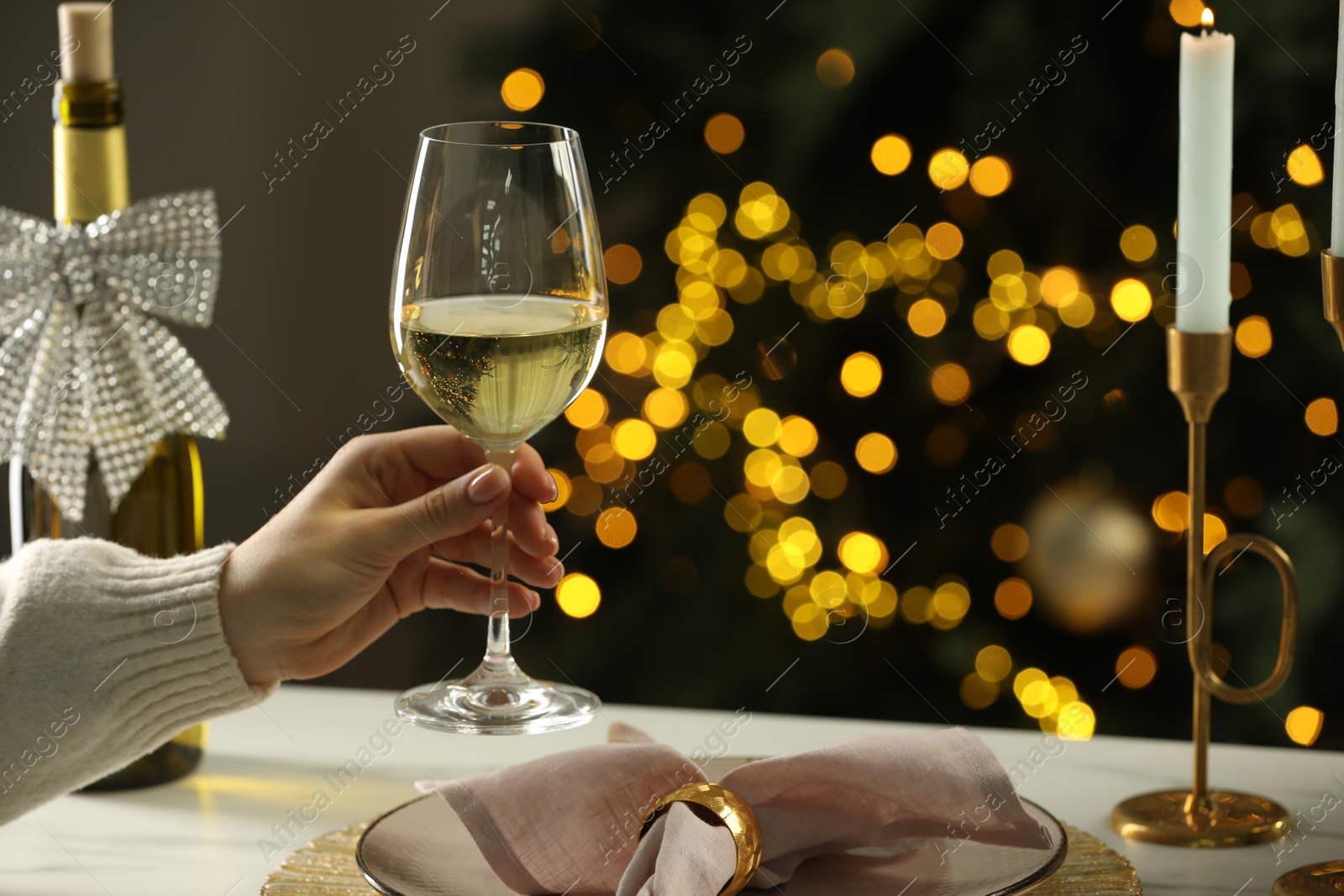 Photo of Woman with glass of white wine at table against blurred Christmas lights, closeup. Bokeh effect