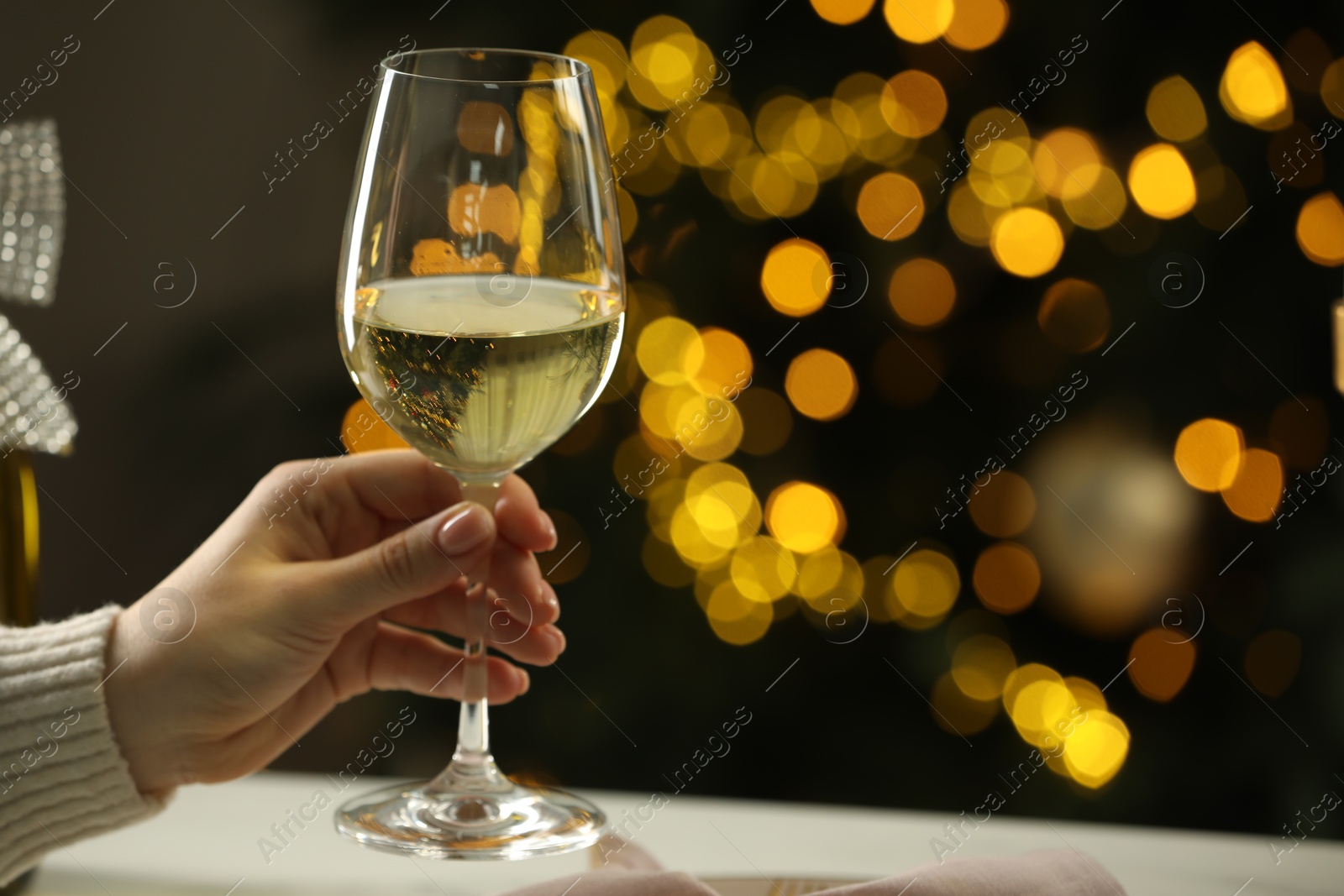 Photo of Woman with glass of white wine at table against blurred Christmas lights, closeup and space for text. Bokeh effect