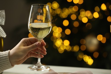 Woman with glass of white wine at table against blurred Christmas lights, closeup and space for text. Bokeh effect