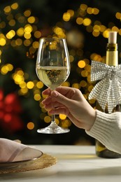 Photo of Woman with glass of white wine at table against blurred Christmas lights, closeup. Bokeh effect