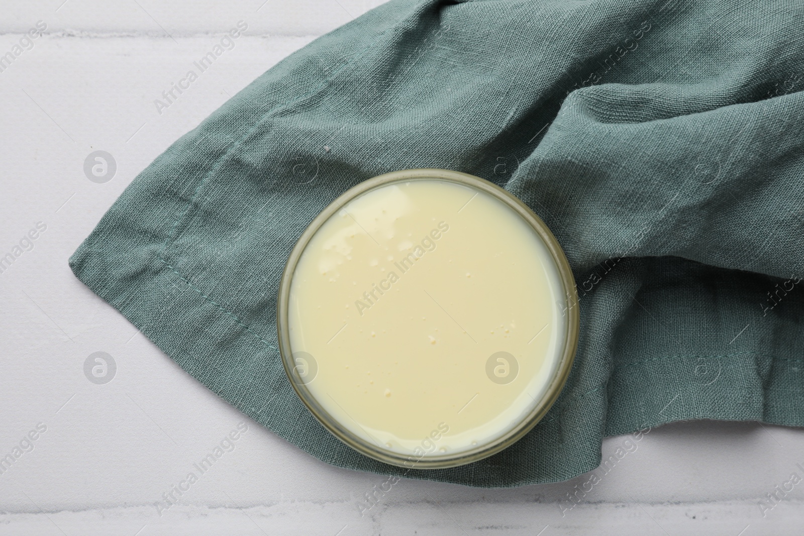 Photo of Tasty condensed milk in bowl and towel on white tiled table, top view