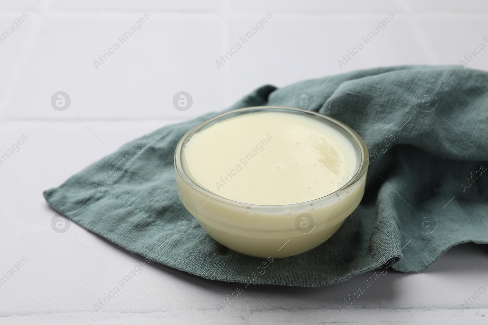 Photo of Tasty condensed milk in bowl and towel on white tiled table, closeup