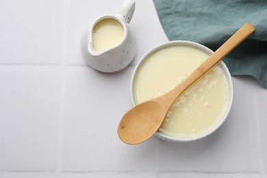 Photo of Tasty condensed milk in bowl and jug with spoon on white tiled table, top view. Space for text