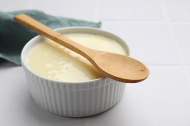 Photo of Tasty condensed milk in bowl and spoon on white tiled table, closeup