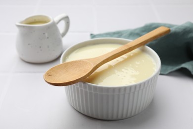 Photo of Tasty condensed milk in bowl and jug with spoon on white tiled table, closeup