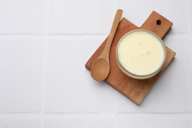 Photo of Tasty condensed milk in bowl and spoon on white tiled table, top view. Space for text