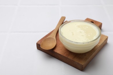 Photo of Tasty condensed milk in bowl and spoon on white tiled table, closeup. Space for text