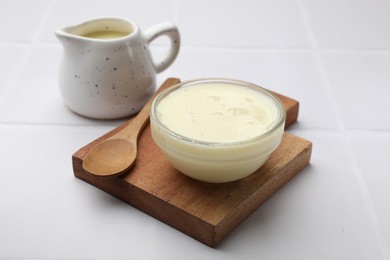 Photo of Tasty condensed milk in bowl and jug with spoon on white tiled table, closeup