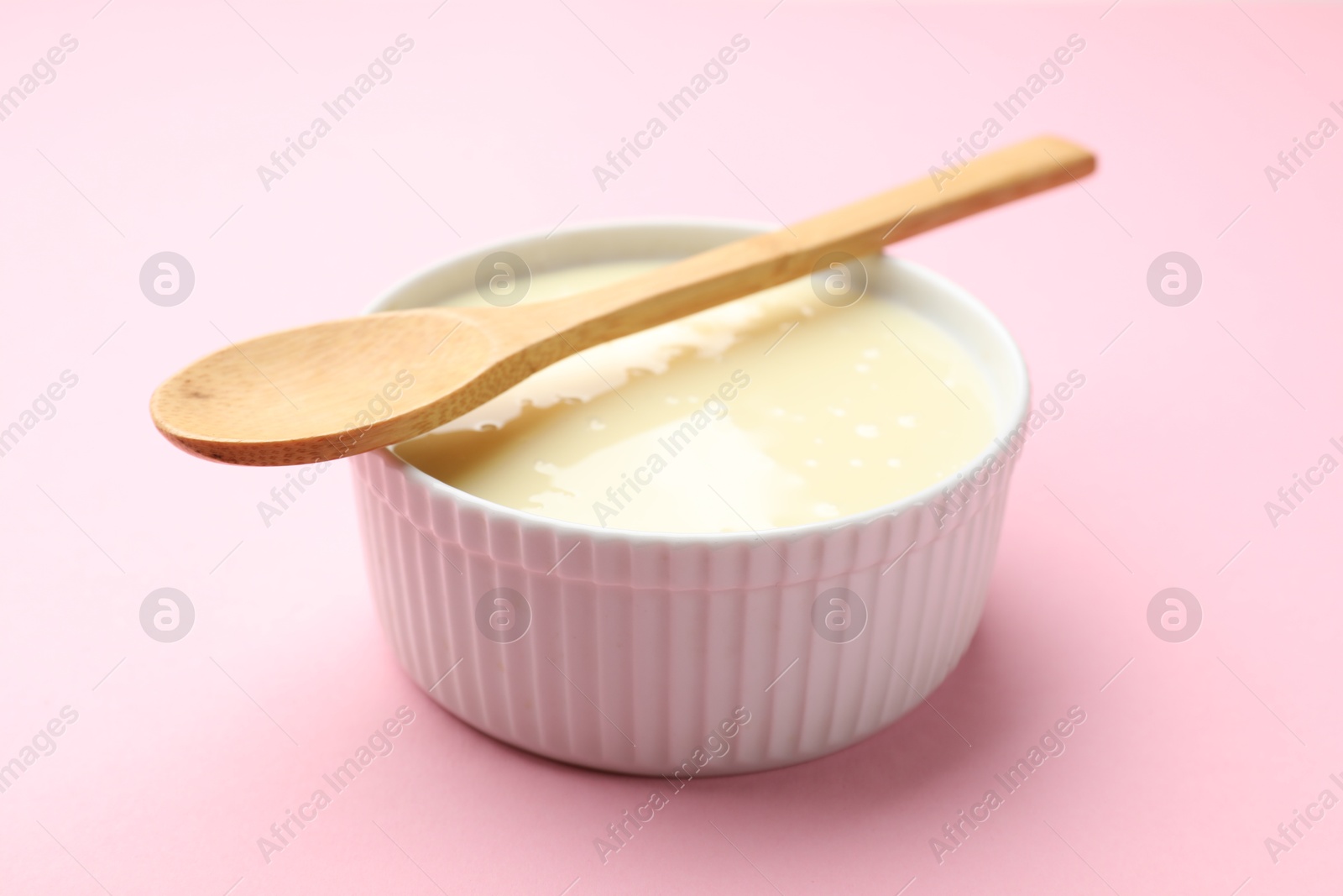 Photo of Tasty condensed milk in bowl and spoon on light pink background, closeup