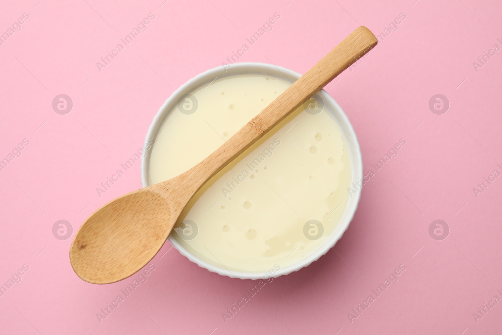 Photo of Tasty condensed milk in bowl and spoon on light pink background, top view