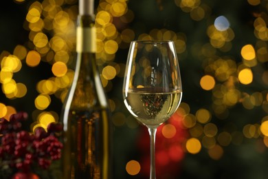 Photo of Bottle and glass of white wine against blurred Christmas lights, closeup. Bokeh effect