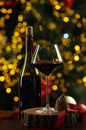 Photo of Glass and bottle of red wine with Christmas decor on wooden table against blurred lights. Bokeh effect