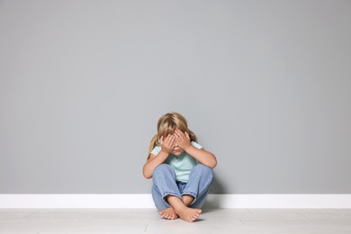 Photo of Orphanage concept. Sad little girl closing her face with hands on floor near grey wall