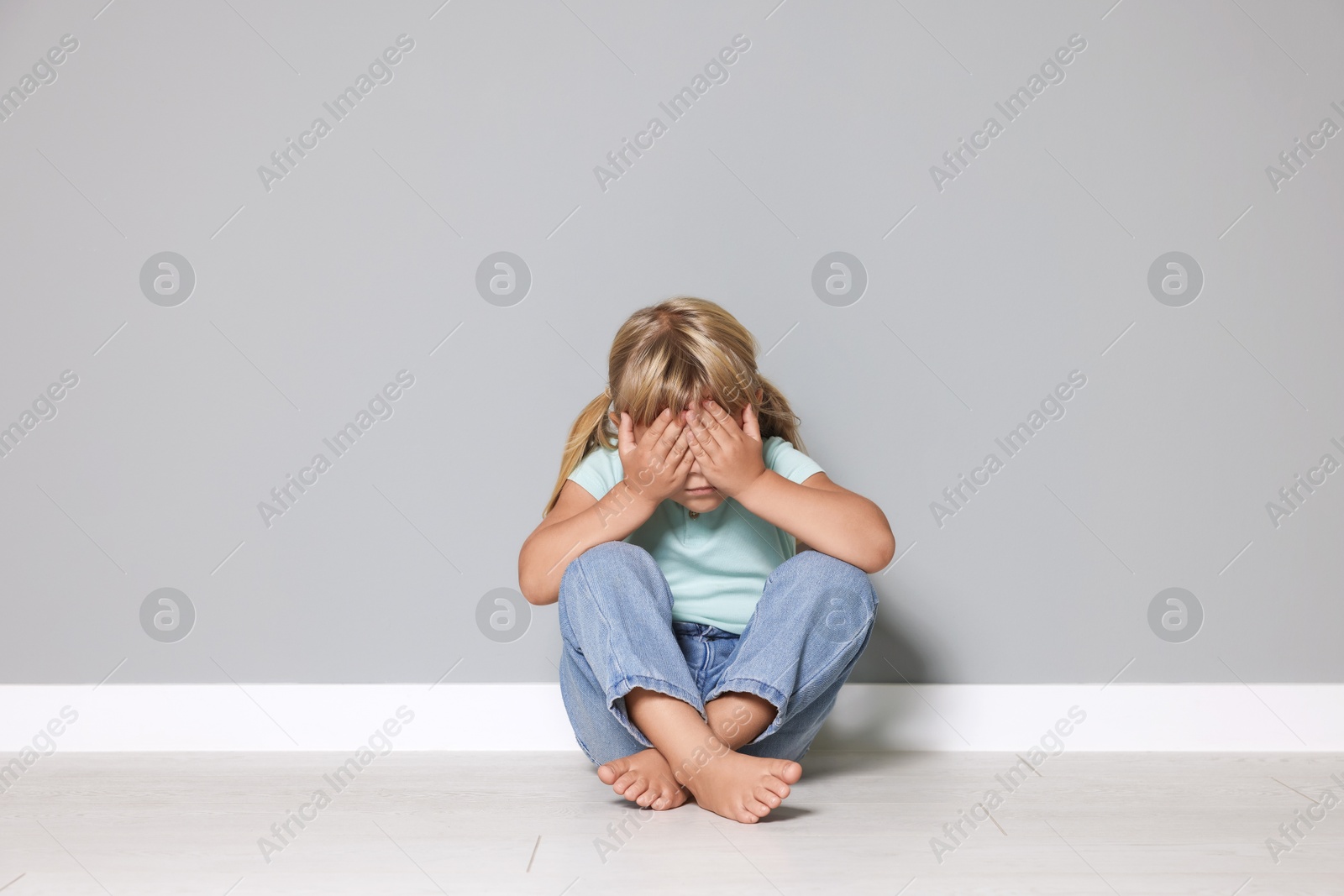 Photo of Orphanage concept. Sad little girl closing her face with hands on floor near grey wall