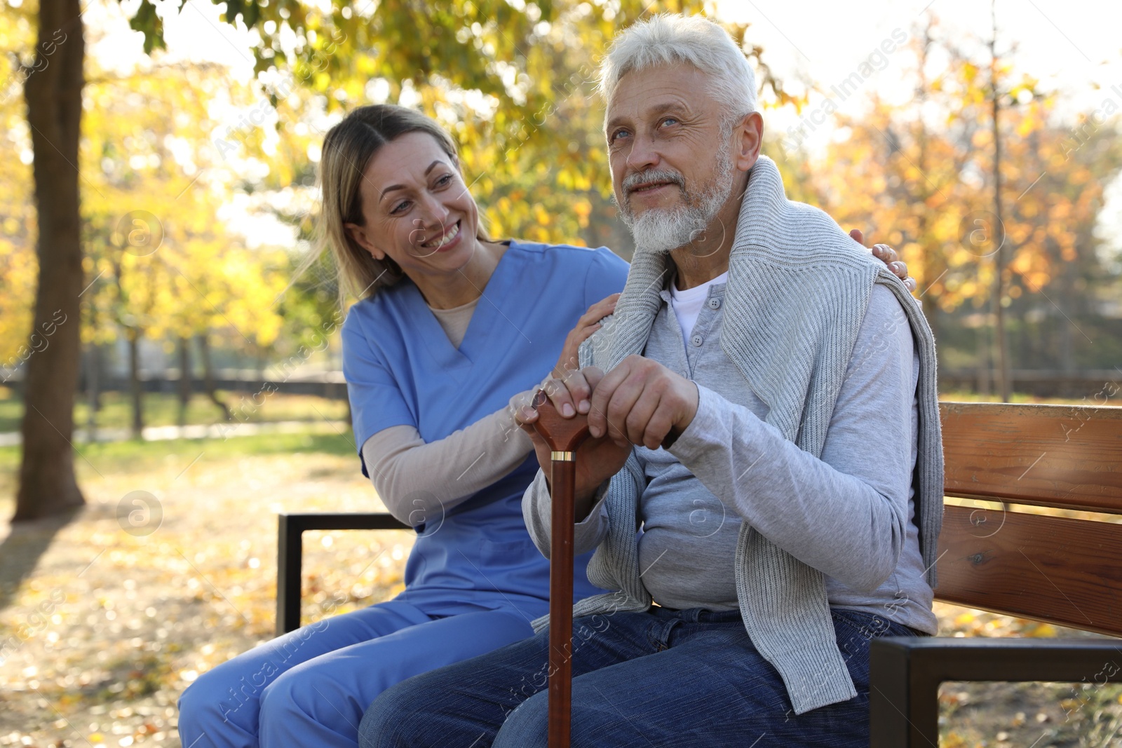 Photo of Caregiver assisting senior man on wooden bench in park. Home health care service