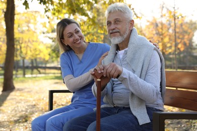 Photo of Caregiver assisting senior man on wooden bench in park. Home health care service