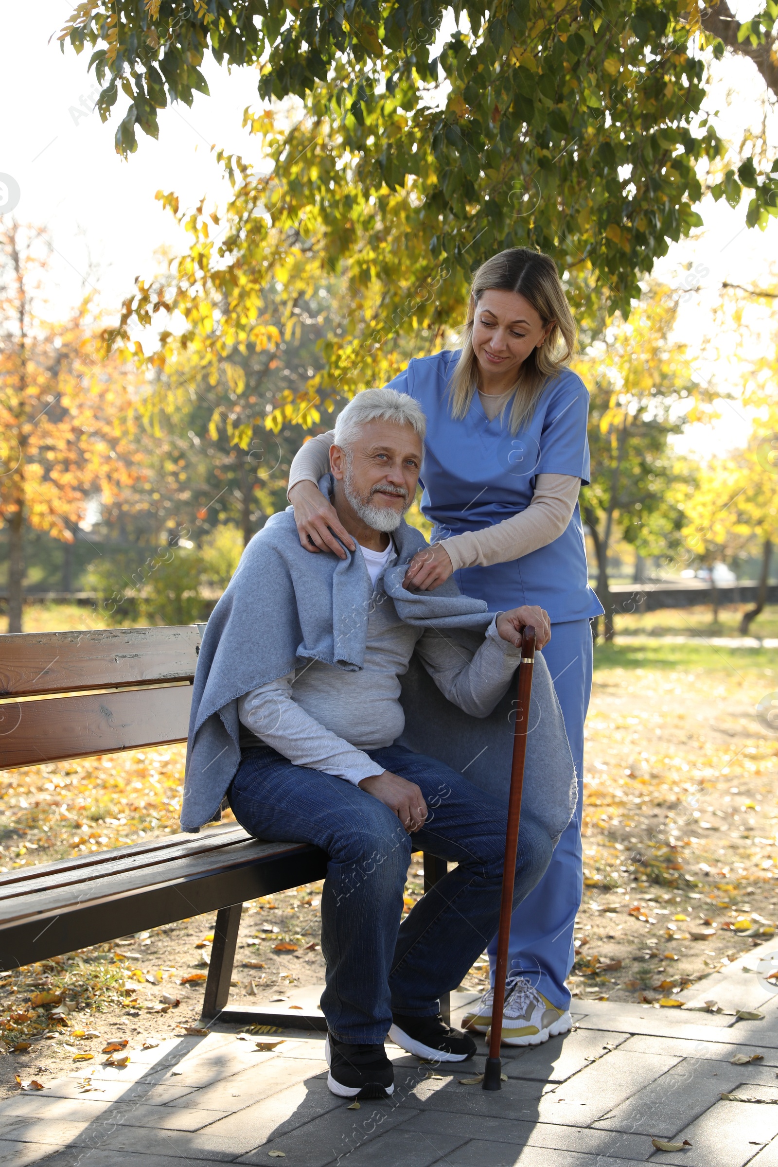 Photo of Caregiver assisting senior man on wooden bench in park. Home health care service