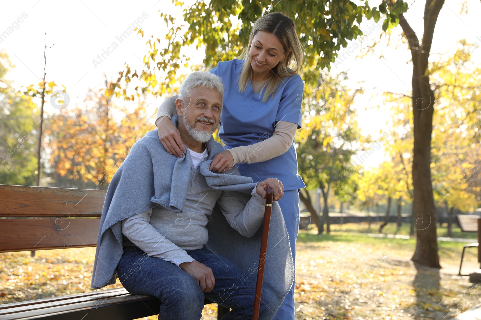 Photo of Caregiver assisting senior man on wooden bench in park. Home health care service