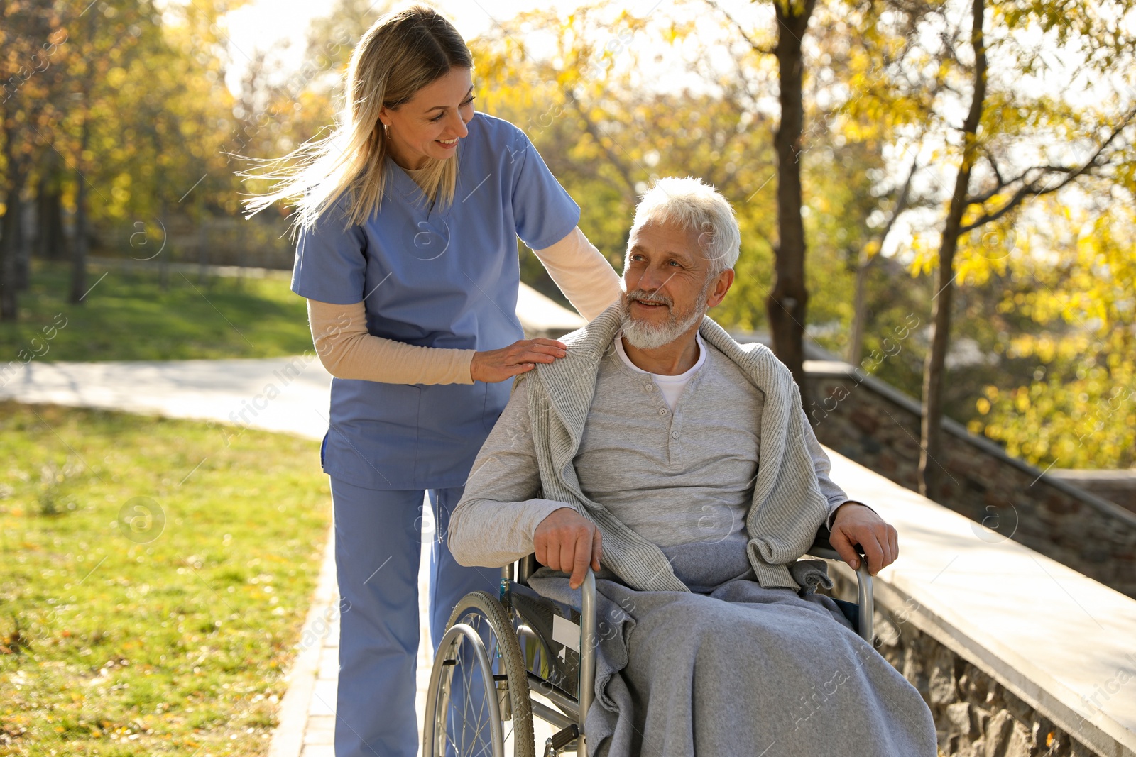 Photo of Caregiver assisting senior man on wheelchair in park. Home health care service