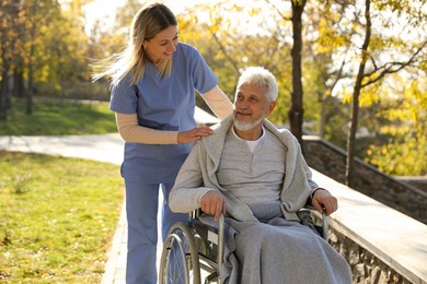 Photo of Caregiver assisting senior man on wheelchair in park. Home health care service