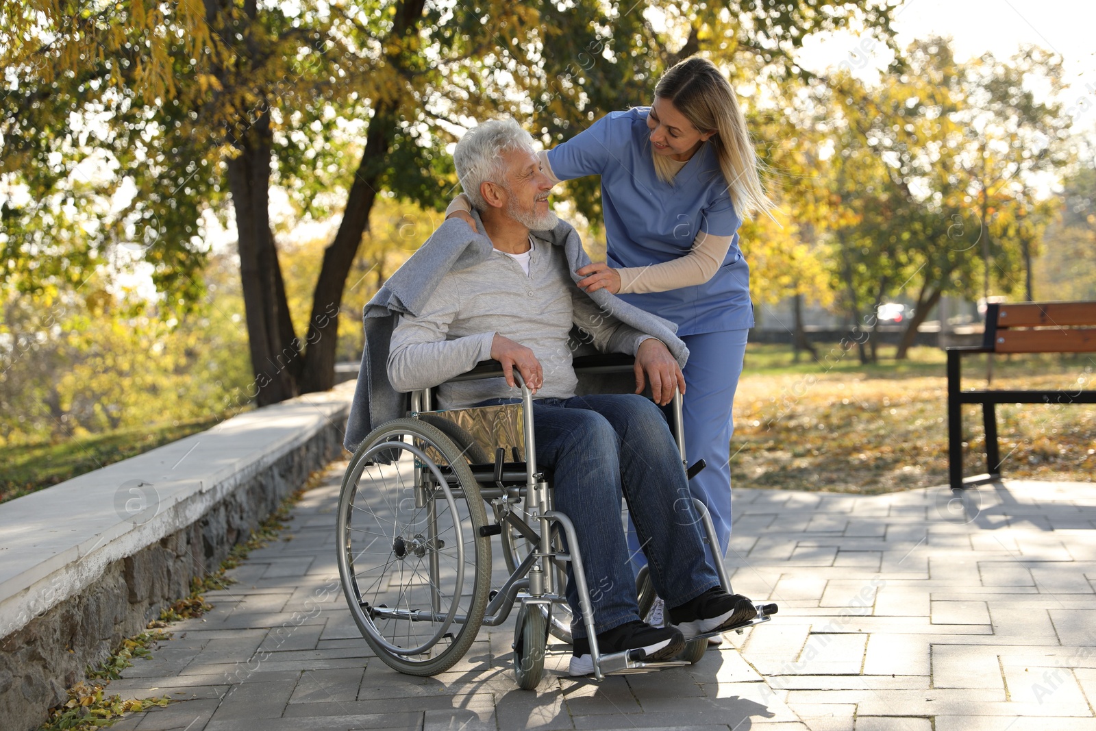 Photo of Caregiver assisting senior man on wheelchair in park. Home health care service