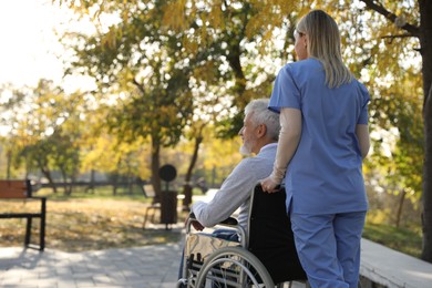 Photo of Caregiver assisting senior man on wheelchair in park, back view and space for text. Home health care service