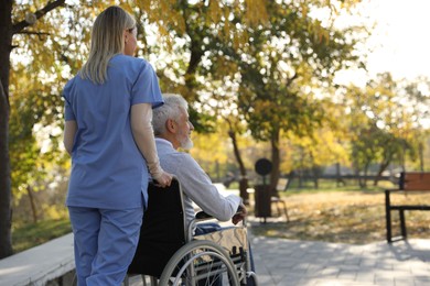 Photo of Caregiver assisting senior man on wheelchair in park, back view and space for text. Home health care service