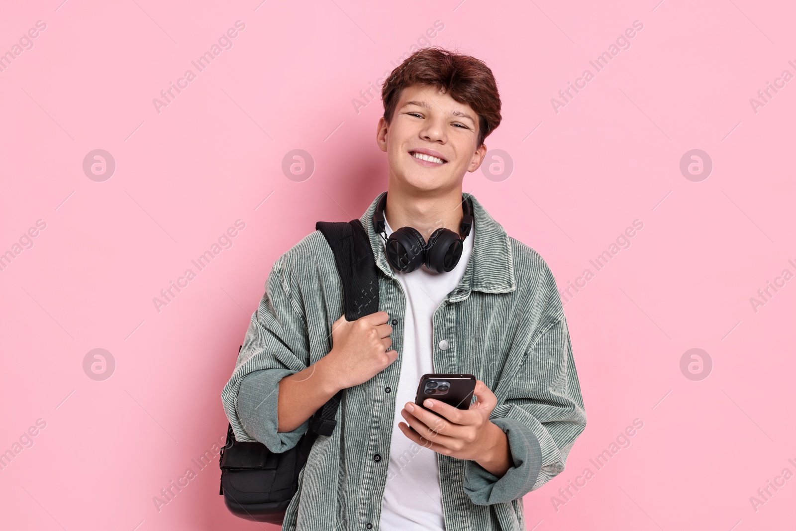 Photo of Happy teenage boy with headphones, smartphone and backpack on pink background