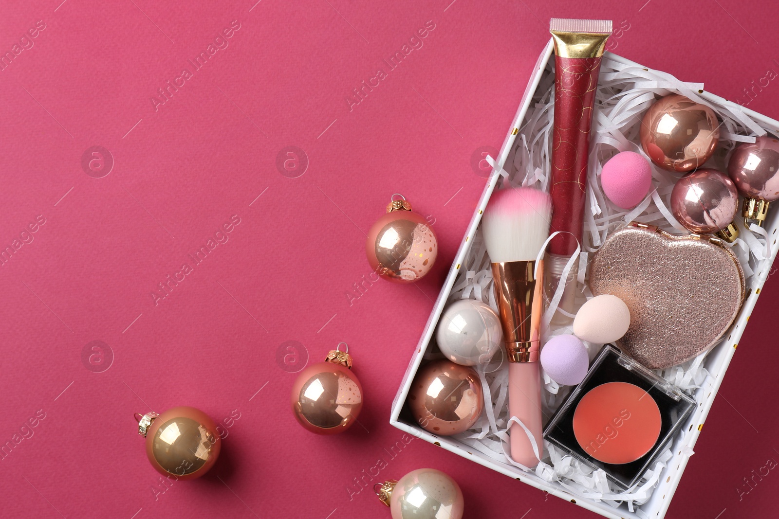 Photo of Christmas present. Decorative cosmetics in gift box and festive balls on dark pink background, top view. Space for text