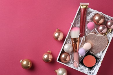 Christmas present. Decorative cosmetics in gift box and festive balls on dark pink background, top view. Space for text