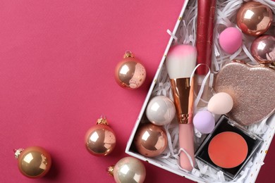 Photo of Christmas present. Decorative cosmetics in gift box and festive balls on dark pink background, top view. Space for text